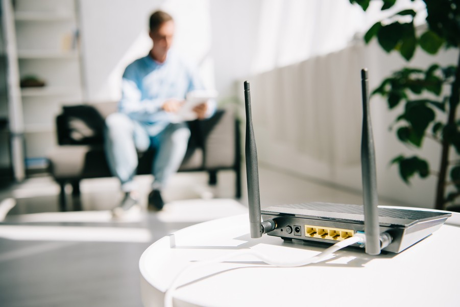 A home networking router in the foreground with a man sitting on a couch in the background. 